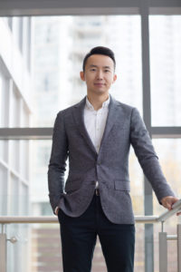 Mark Song is standing by a window in a suit jacket and open-collar shirt. 