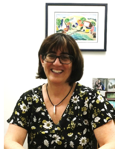 Deborah has dark brown shoulder length hair, glasses, and is wearing a dark flowery shirt. She's in her office smiling at the camera. 