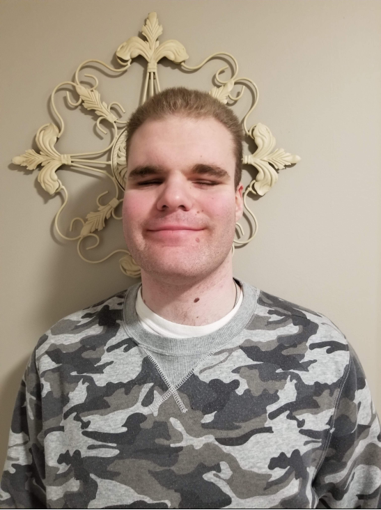 Selfie of Jeffrey. He has dark blonde hair, and is standing against a wall smiling at the camera while wearing a camo shirt.