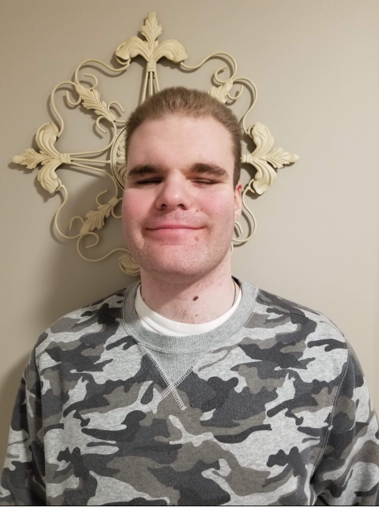 Selfie of Jeffrey. He has dark blonde hair, and is standing against a wall smiling at the camera while wearing a camo shirt. 