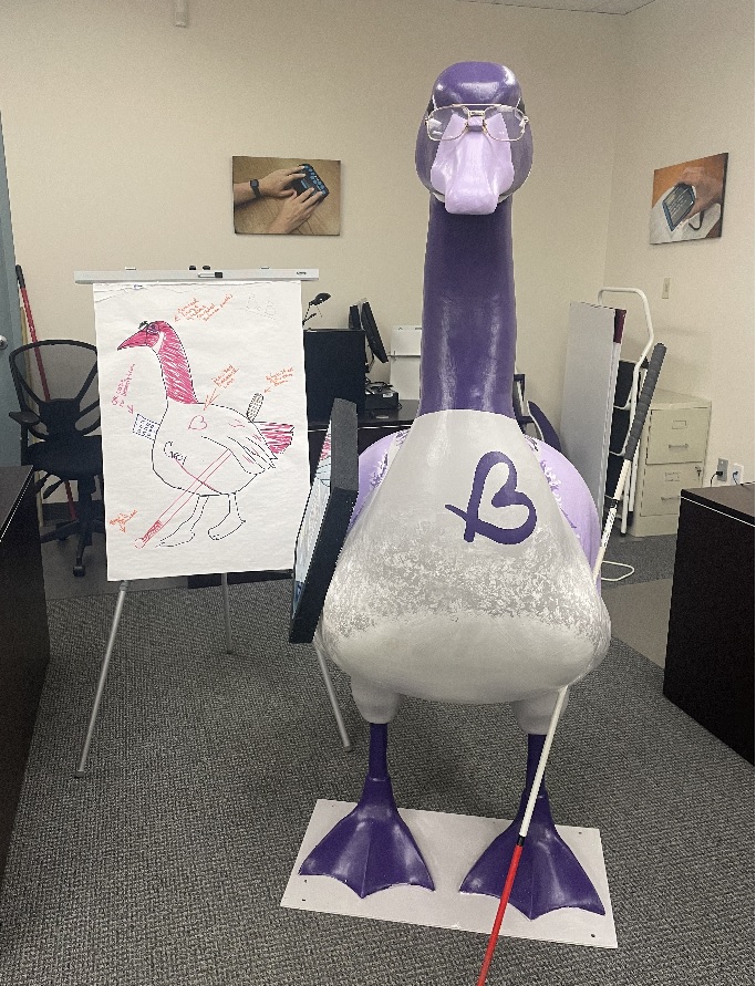 6-ft tall Goose statue, painted in grey with light purple wings and beak, and a darker purple neck. The Goose is wearing eyeglasses; A white cane is attached to it's right wing, and a tablet is attached to it's left wing.
